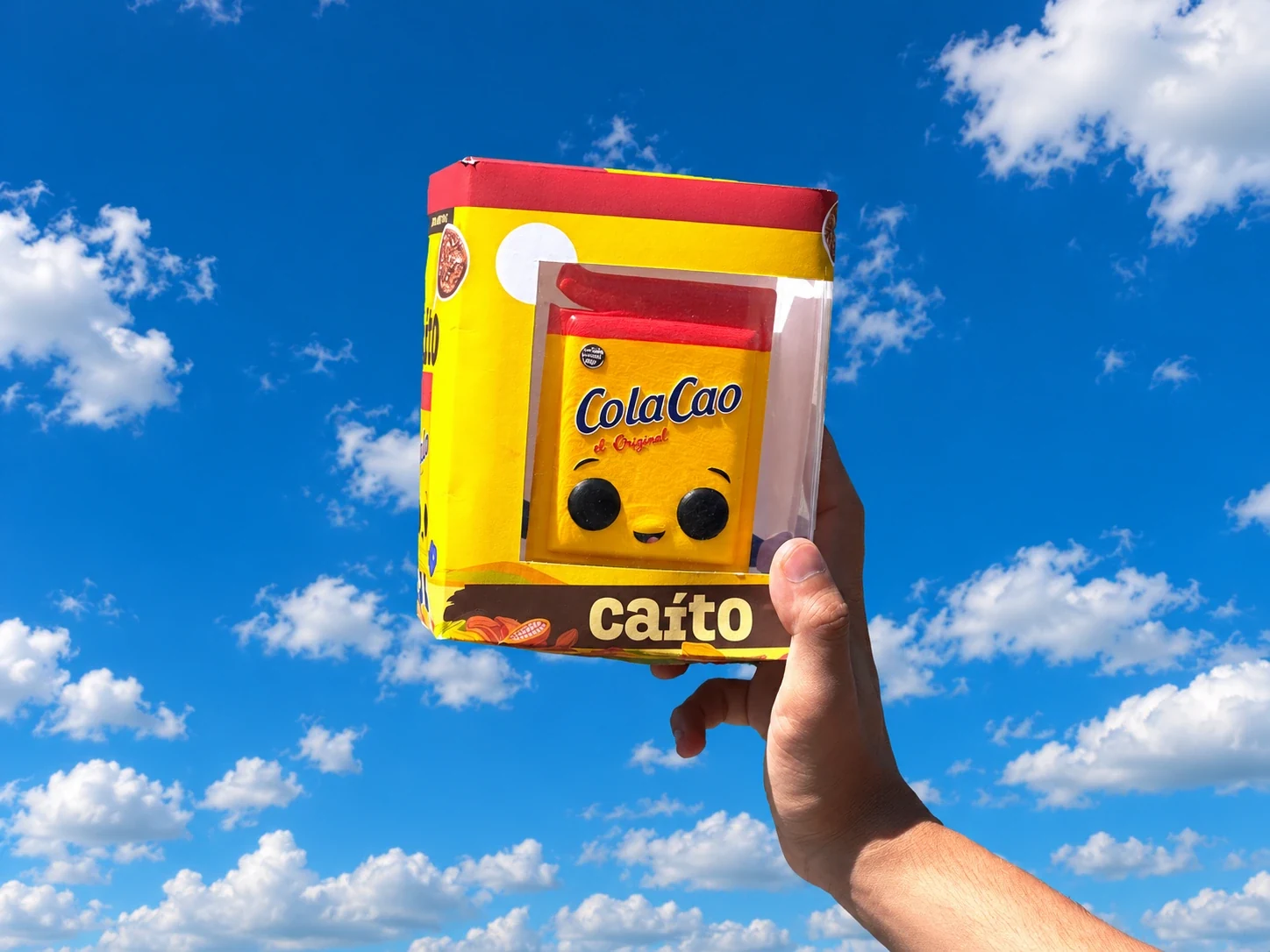 La figura de Caíto en su caja de ColaCao, sostenida contra el cielo azul