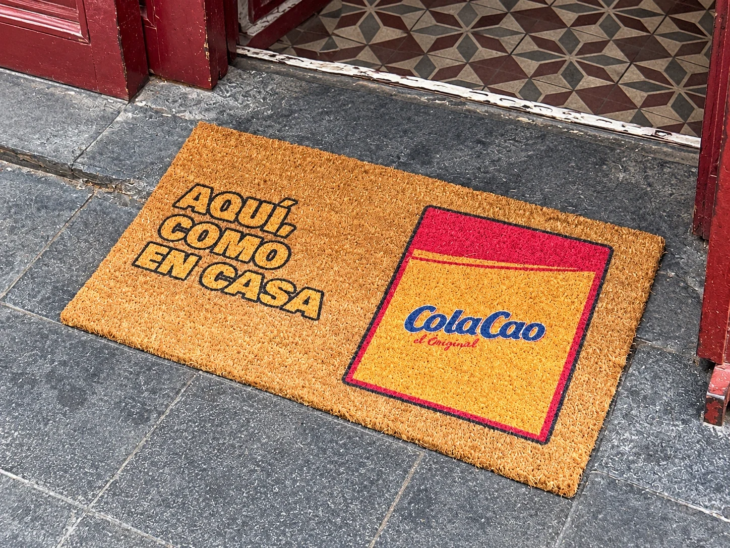 Felpudo de ColaCao con el mensaje Aquí, como en casa en la entrada de un bar